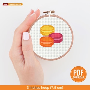 May include: A cross-stitch embroidery hoop featuring a design of three colorful macarons in yellow, orange, and pink. The hoop is 3 inches (7.5 cm) in diameter and is held in a hand. The image includes the text "PDF DOWNLOAD" and the website address "LABPATTERN.COM".