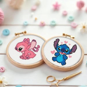 May include: Two cross-stitch embroidery hoops featuring cartoon characters, one pink and one blue, with wooden frames. The pink character has wings, and the blue character has hearts. Gold scissors are also in the image.