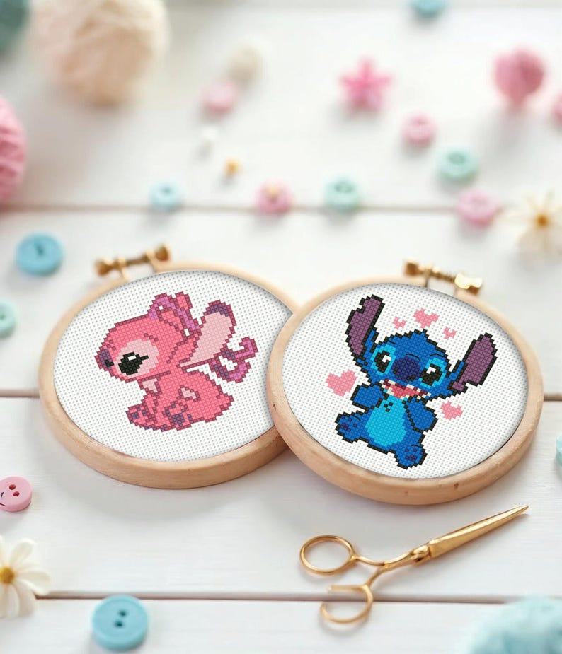 May include: Two cross-stitch embroidery hoops featuring cartoon characters, one pink and one blue, with wooden frames. The pink character has wings, and the blue character has hearts. Gold scissors are also in the image.