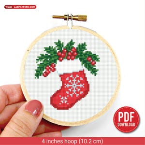 Può includere: Un telaio da ricamo a punto croce con un motivo a calza natalizia rossa con un fiocco di neve, ornato da foglie verdi e bacche rosse. Il telaio in legno misura 10,2 cm di diametro. Il testo "PDF DOWNLOAD" e "www.labpattern.com" sono inclusi.
