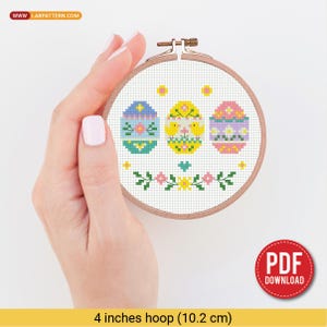 May include: A cross-stitch embroidery hoop featuring three Easter eggs in pastel colors. The eggs are decorated with floral and geometric patterns. The hoop is 4 inches (10.2 cm) in diameter and has a wooden frame. The design includes the text "PDF DOWNLOAD".