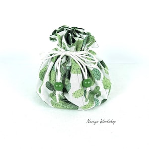 May include: A small, round fabric pouch with a white background and a pattern of green cacti. The pouch is gathered at the top with a white drawstring and green beads. The text "Nancys Workshop" is printed on the bottom right.