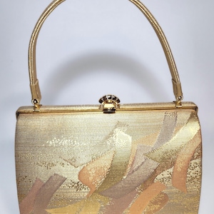May include: A gold-toned handbag with a curved handle and a decorative clasp. The bag features a textured surface with abstract brushstroke patterns in shades of gold, peach, and brown. The handle and frame are also gold.