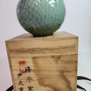 Könnte beinhalten: Eine hellgrüne Keramikvase mit strukturierter Oberfläche, auf einer Holzkiste. Die Vase ist rund mit einer kleinen Öffnung oben. Die Holzkiste hat japanische Schriftzeichen und ein Band. Ungefähr 15 cm hoch.