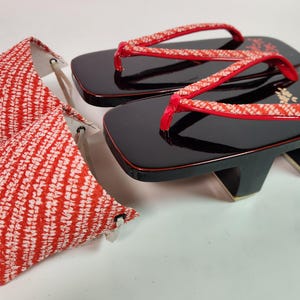 Könnte beinhalten: Schwarze Geta-Sandalen aus Holz mit roten Riemen und floralen Akzenten. Ein passender Stoff mit rotem und weißem Muster ist ebenfalls vorhanden. Die Sandalen haben ein traditionelles japanisches Design.