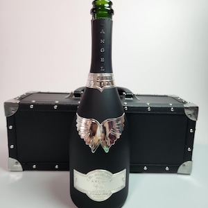 May include: A matte black champagne bottle featuring a silver metallic angel wing emblem on the front, a silver label with the text "CHAMPAGNE ANGEL", and the word "ANGEL" printed vertically on the neck. The bottle is positioned in front of a black case.