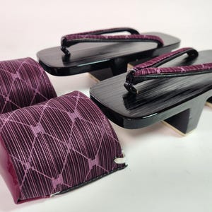 Könnte beinhalten: Paar traditionelle japanische Geta-Sandalen mit schwarzen Holzplattformen und lila Stoffriemen. Die Sandalen werden von zwei passenden lila Stoffbezügen mit einem geometrischen Muster begleitet.