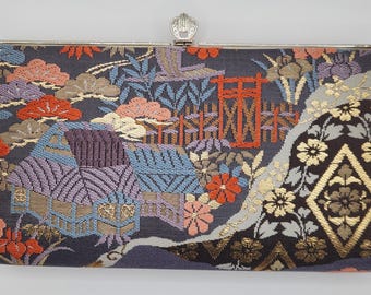 Puede incluir: Bolso de mano rectangular con cierre plateado. El diseño tejido muestra casas, árboles y flores en tonos morados, naranjas, azules y dorados sobre un fondo oscuro. Patrón decorativo.