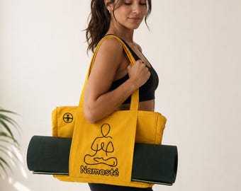 Puede incluir: Un bolso tote de lona color amarillo mostaza con una ilustración de línea negra de una persona en postura de yoga sentada sobre el texto Namasté. El bolso tiene dos correas para el hombro y una esterilla de yoga verde oscuro horizontal.