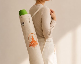 May include: A cream-colored yoga mat bag with a shoulder strap, carrying a rolled-up green yoga mat. The bag features an orange illustration of a seated figure within a lotus flower design. The bag is approximately 30 inches long.