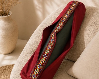 May include: A burgundy yoga mat bag with a colorful patterned trim. The bag is open, revealing a dark green yoga mat inside. The bag is made of a soft fabric and is designed to carry a yoga mat. The bag is sitting on a beige chair.