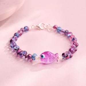 May include: A beaded bracelet featuring a central purple glass fish charm with a blue and white eye detail. The bracelet is composed of translucent purple and blue glass beads interspersed with small white beads and a silver-toned metal clasp.