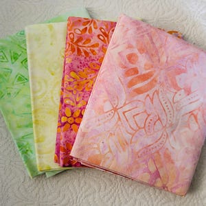 May include: A stack of colorful fabric squares in shades of green, yellow, pink, and orange. Each fabric square features a unique pattern, including geometric shapes and floral designs. The fabrics are folded and stacked, showcasing their vibrant colors and textures.