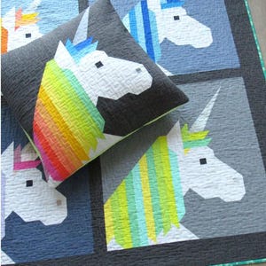 May include: A square pillow with a unicorn design rests on a quilt with similar unicorn patterns. The pillow features a white unicorn head with a rainbow mane and a dark gray background. The quilt has a grid pattern with various unicorn heads.
