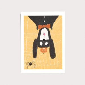 Peut inclure: Une impression d'art rectangulaire repr&eacute;sentant un chien noir avec un poitrail blanc et un n&oelig;ud papillon orange suspendu &agrave; l'envers sur un fond jaune avec un motif de toile d'araign&eacute;e blanche. Une petite araign&eacute;e noire et une citrouille sont visibles.