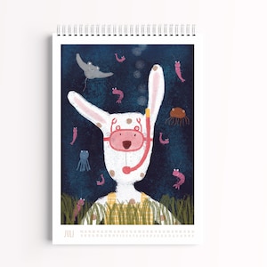 Peut inclure: Un calendrier mural &agrave; spirale pour le mois de juillet illustr&eacute; d'un lapin blanc portant un masque de plong&eacute;e rose et une salopette jaune. Le fond repr&eacute;sente une sc&egrave;ne sous-marine bleu fonc&eacute; avec une raie, des petites crevettes roses et une m&eacute;duse.