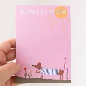 Puede incluir: Tarjeta rosa con el texto "HEUT MACH ICH'S MIR SCHÖN" y una ilustración de un perro salchicha con una camisa a rayas y una boina, sosteniendo un helado. Un sol amarillo está en la esquina superior derecha.