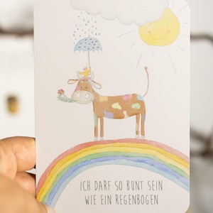 Peut inclure: Illustration à l'aquarelle d'une vache sur un arc-en-ciel. La vache a des taches colorées, une couronne et tient une fleur. Un soleil et un nuage de pluie sont au-dessus de la vache. Le texte dit : "Je peux être aussi coloré qu'un arc-en-ciel."