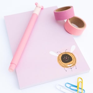 Könnte beinhalten: Ein hellrosa Notizblock mit einer Cartoon-Bienenillustration in der unteren rechten Ecke, begleitet von einem passenden rosa Stift mit Figurenaufsatz, zwei Rollen rosa Washi-Tape und drei bunten Büroklammern in Weiß, Blau und Gelb.