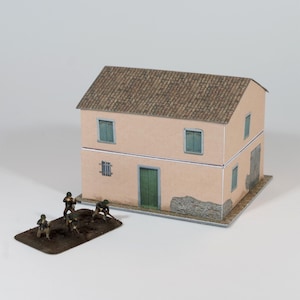 Può includere: Un modello in miniatura di un edificio a due piani con tetto in tegole marroni e pareti color pesca. L'edificio ha persiane verdi e una porta verde. Piccoli soldatini sono posizionati di fronte all'edificio.