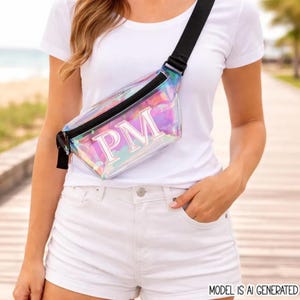 May include: A clear iridescent fanny pack with a black zipper and strap. The bag features the white capital letters PM printed on the front. The material reflects various colors including pink, blue, and purple, creating a holographic effect.