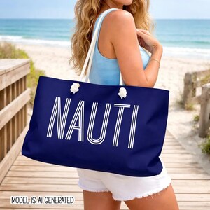 May include: A large navy blue tote bag featuring the word NAUTI in bold white capital letters with a striped design. The bag has thick white rope handles attached through silver-toned metal grommets, providing a sturdy and functional carry option.