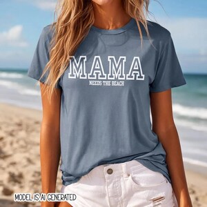 May include: A slate blue short-sleeved t-shirt featuring the white text MAMA in a collegiate font, with the words NEEDS THE BEACH printed in a smaller white sans-serif font directly underneath the main text on the front of the garment.