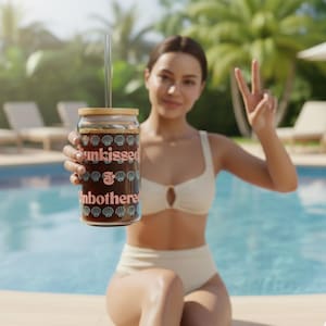 May include: A clear glass tumbler with a bamboo lid and a straw, filled with a dark beverage. The tumbler features the words "unkissed & unbothered" in pink and blue, with seashell graphics. The person is wearing a white bikini.
