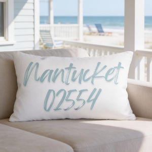 May include: White rectangular pillow with the word "Nantucket" and the number "02554" printed in light blue. The pillow is on a beige sofa, with a beach scene in the background.