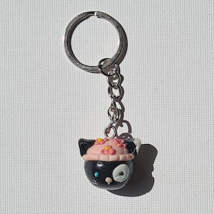 Op de afbeelding: Een zwart en roze sleutelhanger met een katachtig ontwerp, met een roze hoed versierd met kleine roze bloemen. De hanger is bevestigd aan een zilveren sleutelring en ketting. De kat heeft een wit oog en een blauwe stip.