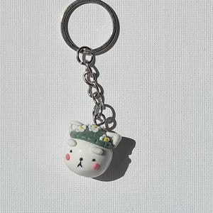 Op de afbeelding: Een witte sleutelhanger in de vorm van een beer met een zilveren ketting en ring. De beer heeft een groene hoed met witte bloemen, roze wangen en zwarte ogen. De sleutelhanger staat op een witte, getextureerde achtergrond.
