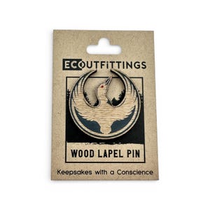 May include: A wooden lapel pin featuring a stylized bird emblem with a red eye, set against a light wood grain. The pin is mounted on a brown card with the text "ECOUTFITTINGS" and "WOOD LAPEL PIN". The card also reads "Keepsakes with a Conscience".