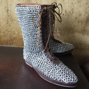 Puede incluir: Un par de botas de cota de malla de estilo medieval. Las botas tienen una parte superior de cota de malla plateada, detalles de cuero marrón y cordones. Las suelas son de color marrón oscuro. Las botas se exhiben sobre una superficie marrón oscuro.