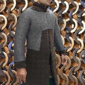 May include: A silver chainmail shirt over a brown padded garment. The chainmail covers the upper body, while the padded garment extends to the knees. The sleeves are long, and the collar is high. The background is a pattern of metal links.