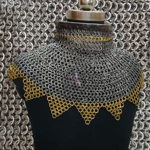 May include: A chainmail coif, a medieval neck and shoulder defense, is displayed on a black mannequin. The coif features a silver chainmail body with gold-colored trim along the neckline and lower edge. The background is a chainmail pattern.