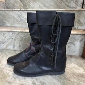 May include: A pair of black leather mid-calf boots featuring a folded cuff at the top and a vertical lace-up closure on the outer side with matching black leather laces. The boots have a rounded toe, a flat sole, and a smooth, matte finish throughout.