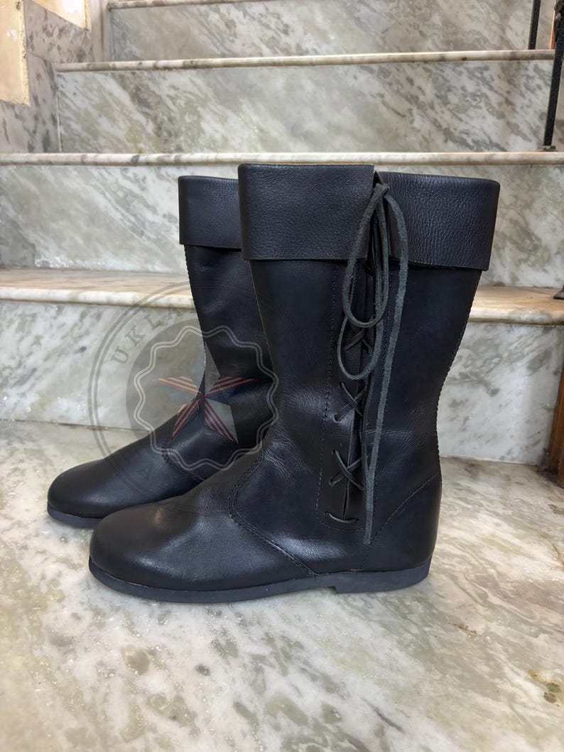 May include: A pair of black leather mid-calf boots featuring a folded cuff at the top and a vertical lace-up closure on the outer side with matching black leather laces. The boots have a rounded toe, a flat sole, and a smooth, matte finish throughout.