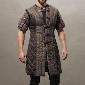 Könnte beinhalten: Brauner, gesteppter mittelalterlicher Gambeson mit kurzen Ärmeln, Lederdetails und Metallschnallen. Das lange, tunikaartige Kleidungsstück weist ein Rautenmuster auf und eignet sich für historische Nachstellungen oder Cosplay.