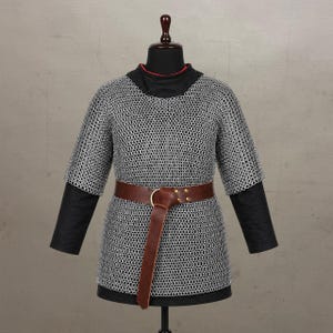 May include: A silver-coloured chainmail hauberk featuring a short-sleeved design with interlocking metal rings. The garment is worn over a black long-sleeved base layer and is cinched at the waist with a wide brown leather belt featuring a metal buckle.