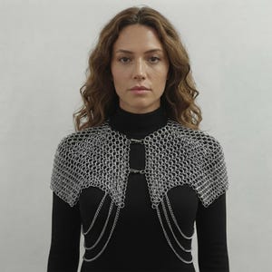 May include: A silver-toned chainmail shoulder piece featuring interlocking metal rings that form a capelet structure. The garment is secured at the front with two metal clasps and includes three draped silver chains hanging from each side.