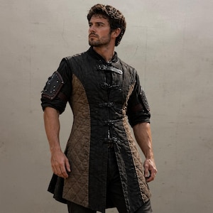 May include: A black and brown quilted medieval-style tunic with leather accents. The tunic features a high collar, decorative buckles, and padded shoulder details. The garment has a tailored fit and a flared hemline, suitable for cosplay or historical reenactment.