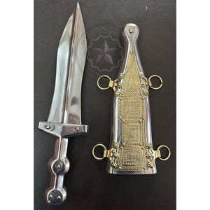 Puede incluir: Una daga plateada con una hoja puntiaguda y un mango decorativo, junto a su vaina. La vaina es plateada con filigrana dorada y cuatro anillos. La daga y la vaina se exhiben sobre una superficie oscura.