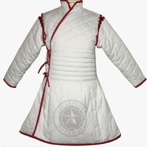 May include: White quilted gambeson with red trim and accents. The medieval garment features long sleeves, a high collar, and a wrap-around design with tie closures. The hemline is flared, and the garment is likely for historical reenactment or costume.