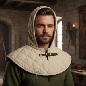 Puede incluir: Una capucha y cuello acolchados de color crema, con una correa de cuero marrón y una hebilla de latón. La capucha tiene un patrón de diamantes y se usa sobre una prenda verde. El diseño recuerda a la vestimenta medieval.
