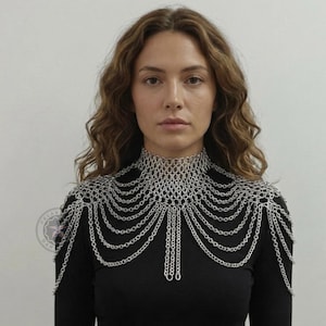 May include: A silver chainmail shoulder piece with a high collar and cascading chains. The intricate design features a woven collar and multiple strands of delicate chains, creating a layered effect. The piece is worn over a black top.