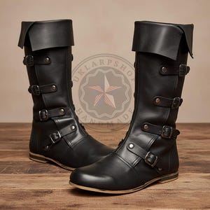 May include: A pair of black knee-high boots featuring a fold-over cuff at the top and four horizontal straps across the front, each secured with a metal buckle and rivets. The boots have a rounded toe and a flat sole with a visible light-coloured edge.