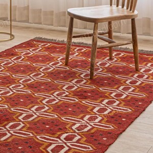 Puede incluir: Una alfombra roja con un diseño geométrico blanco y amarillo se muestra sobre un suelo de madera clara. Una silla de madera está sobre la alfombra. La alfombra tiene flecos en los bordes. Un sofá y una mesa auxiliar están en el fondo.