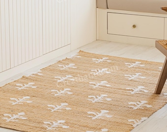 May include: A rectangular jute rug with a repeating white floral pattern. The rug is placed on a light-colored wooden floor in a well-lit room. A wooden chair and a potted plant are also visible.