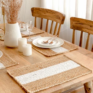 Könnte beinhalten: Ein Esstisch mit gewebten Jute-Tischsets mit einem zentralen weißen Streifen. Der Tisch ist mit weißen Tellern, Besteck, Kerzen und einer weißen Vase mit getrockneten Blumen gedeckt. Holzstühle umgeben den Tisch.