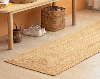 Puede incluir: Una alfombra rectangular de yute con un diseño tejido, colocada sobre un suelo de madera clara. La alfombra es de color beige natural y tiene un borde definido. Un banco de madera, una cesta, zapatos y otros artículos están en el fondo.